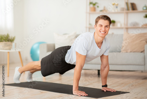 Workout fitness at home. Happy muscular guy doing plank on mat on floor in morning living room interior in morning