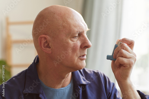 Warm-toned side view portrait of bald senior man using inhaler for asthma or breathing problems in home interior