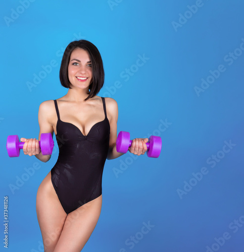 A toned girl in a black sports swimsuit holds dumbbells in her hands  her arms bent at the elbows during exercise on a blue background. Girl is standing with dumbbells. Copy space