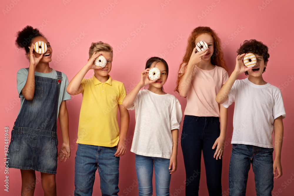 Diverse schoolkids holding yummy donuts in front of their faces over pink background