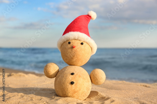 Snowman made of sand with Santa hat on beach near sea. Christmas vacation