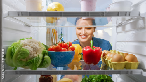 Beautiful Young Woman Looks inside the Fridge and Takes out Vegetables. Woman Preparing Healthy Meal Using Groceries. Point of View POV from Inside of the Kitchen Refrigerator full of Healthy Food