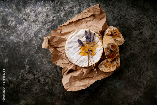 French cheese Camembert covered with honey, dried lavender stems on the paper and freshly baked baguette over dark rustic background. Top view. Copy space