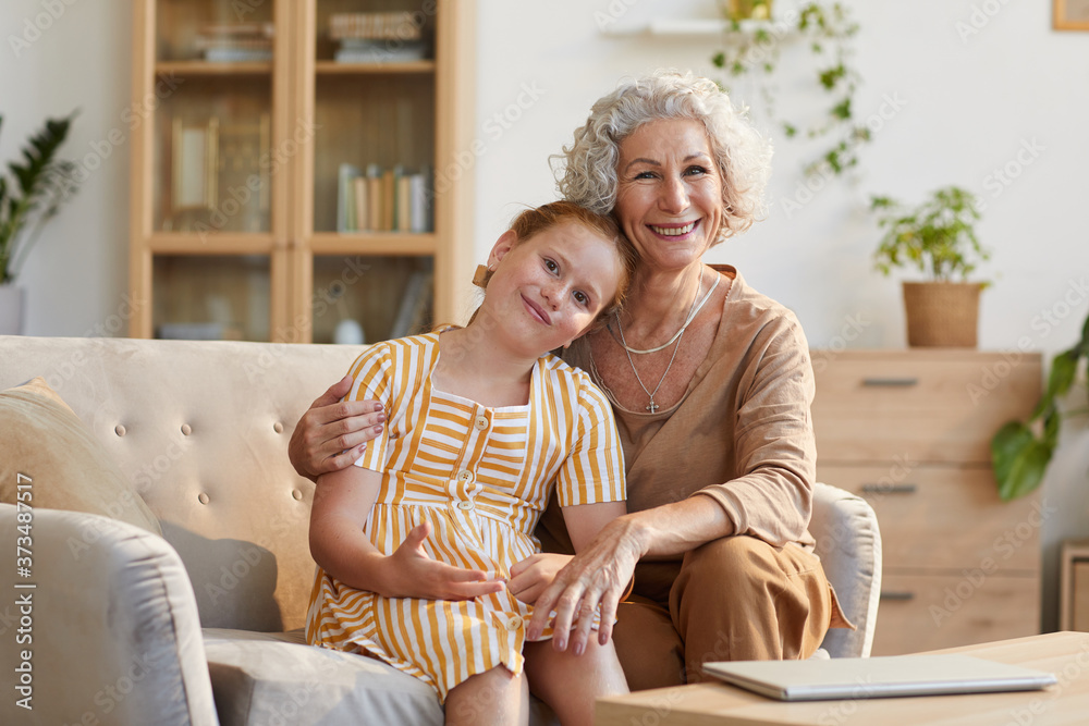 Obraz premium Warm toned portrait of smiling senior woman embracing cute granddaughter while sitting on couch and looking at camera in cozy home interior, copy space