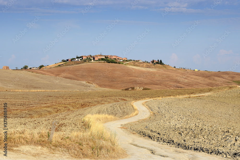 Weg durch die Felder zu einer Siedlung auf einem Hügel in der Crete Senesi Weg durch die Felder zu einer Siedlung auf einem Hügel in der Crete Senesi