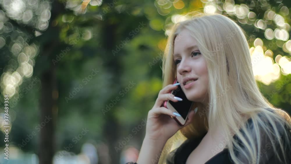 Cute blonde caucasian pretty girl with long hair smiling talking on cell phone in park and hanging up. Concept of relax remote communication with friends, relatives and loved. Ordering by smartphone Cute blonde caucasian pretty girl with long hair smiling talking on cell phone in park and hanging up. Concept of relax remote communication with friends, relatives and loved. Ordering by smartphone