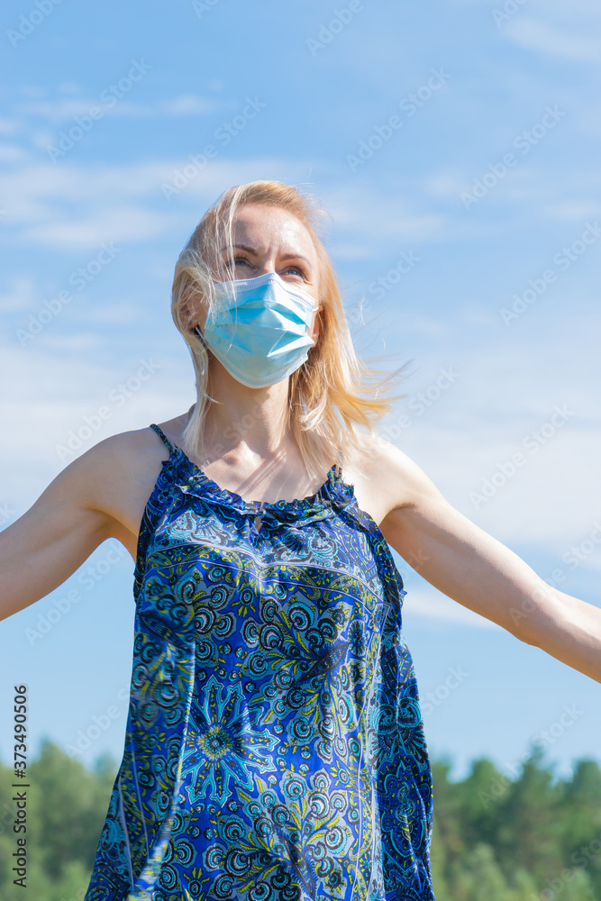 Healthcare and medicine concept. Portrait of blonde woman with windswept hair and medicine mask on her face. Woman wear blue dress in blue sky and green trees background with copy space