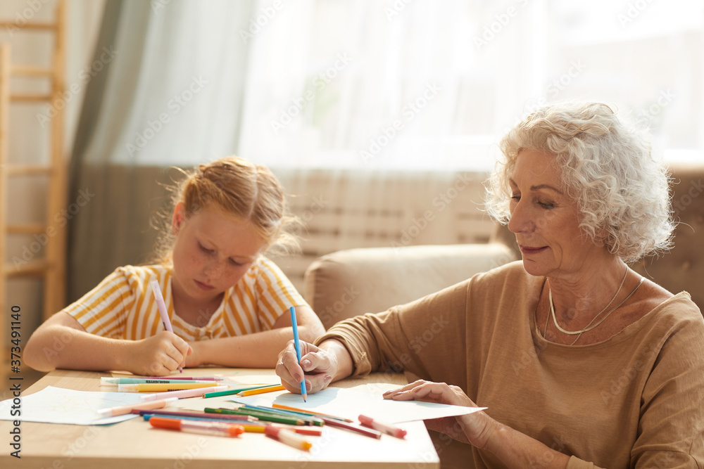 Warm toned portrait of smiling senior woman babysitting cute red haired girl and drawing together while sitting by coffee table in cozy living room Warm toned portrait of smiling senior woman babysitting cute red haired girl and drawing together while sitting by coffee table in cozy living room
