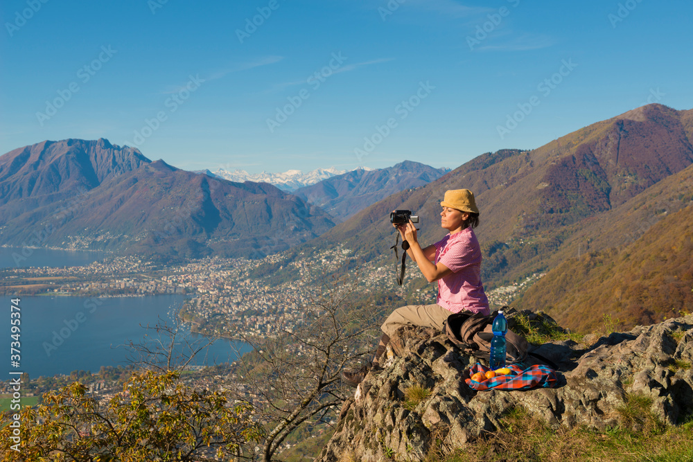 Woman with Hat Sitting on a Mountain Peak and Taking Photos with Her Camera over Alpine Lake Maggiore and Mountain in Ticino, Switzerland.