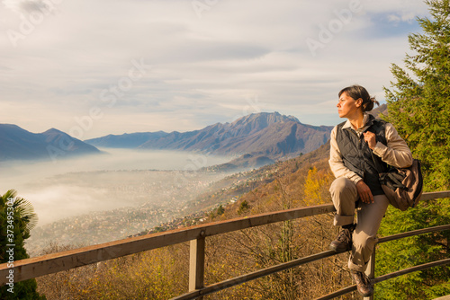 Woman with Backpack Sitting on a Railing and Joy the Panoramic View over an Alpine Lake Maggiore and Mountain with Fog in Ticino, Switzerland.