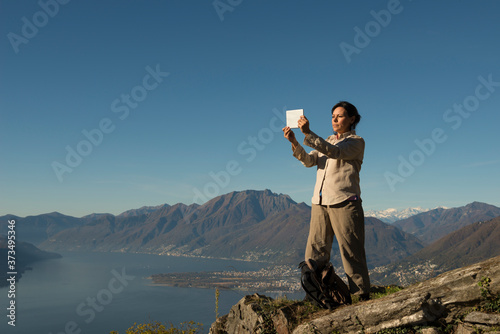 Woman Standing on a Mountain Peak and Taking Photos with Her Tablet over Alpine Lake Maggiore and Mountain in Ticino, Switzerland.