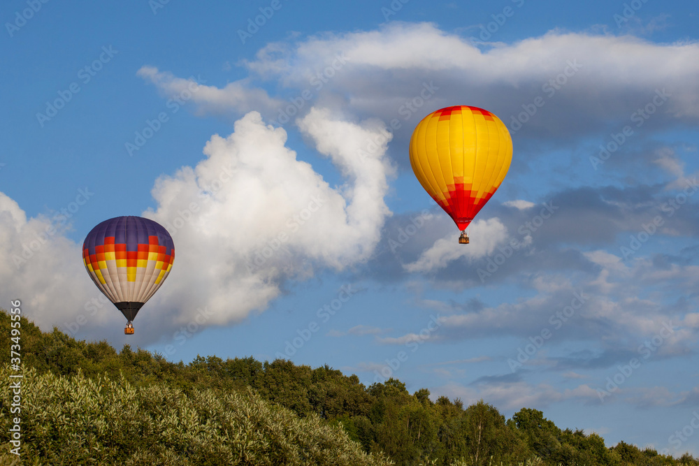 Fototapeta premium Two multi-colored hot air balloons flying over the summer forest. Some clouds in the sky.