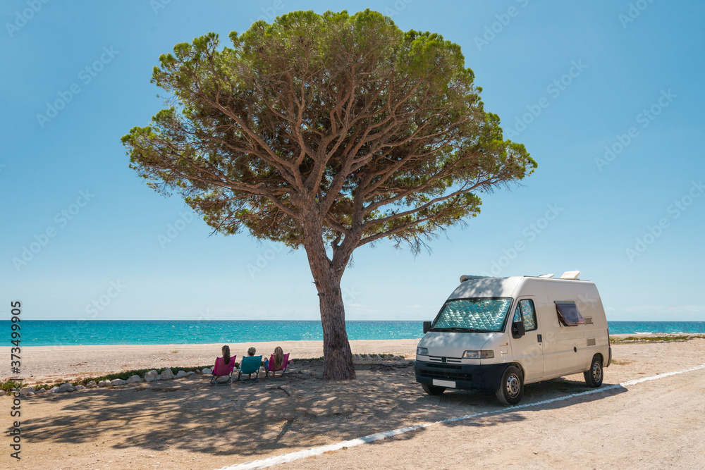 Women sitting in hammocks looking at the beach next to a camper van. They are in the shade of a pine tree. Van life