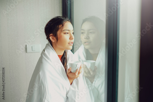 Beautiful asian woman standing by the window, holding coffee in her hand in the morning.