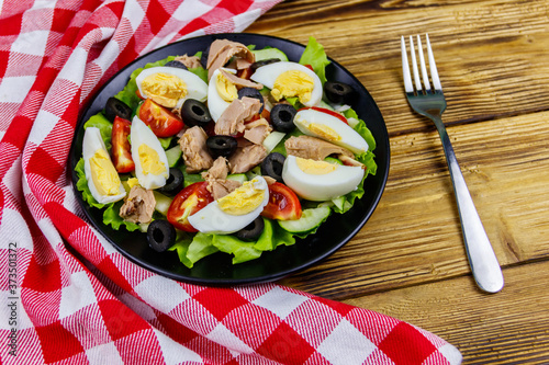 Tasty tuna salad with lettuce, black olives, eggs and fresh vegetables on wooden table