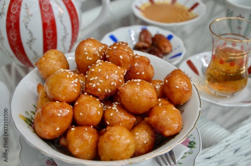 Traditional oriental dessert speciality Legematt (Luqaimat), fried dough balls in a porcelain bowl, blurred dates and a cup of tea behind, blurred background
