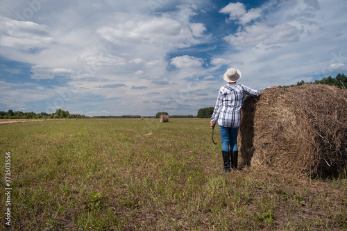 A female farmer stands by a haystack after harvesting in a field with the sky and forest in the background. Landscape