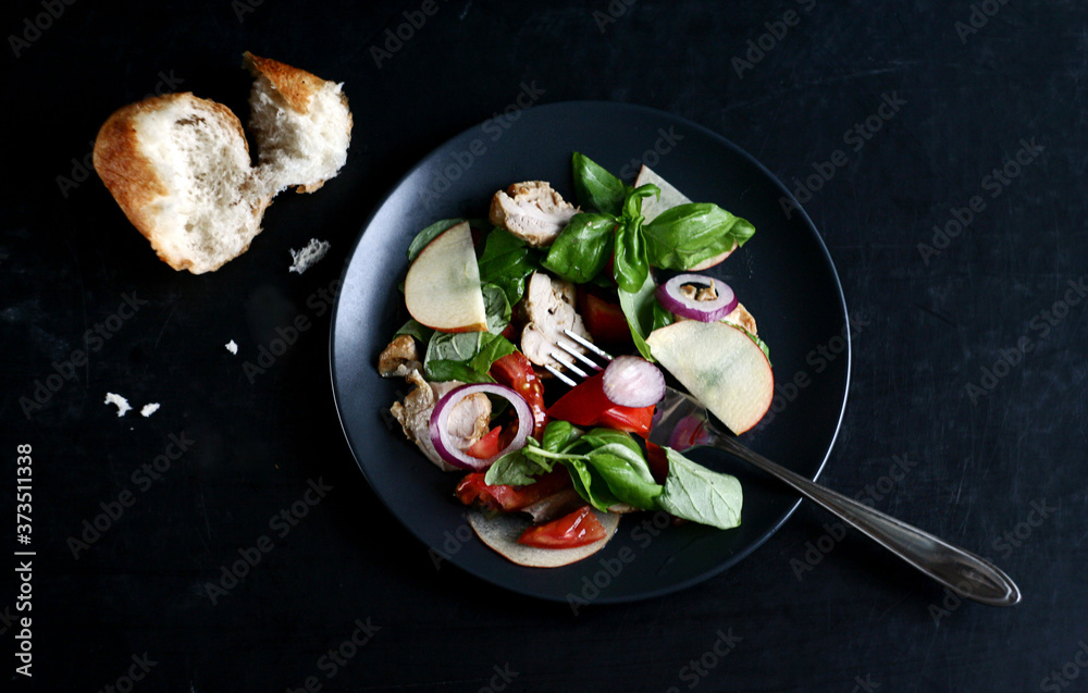 Salad with tomatoes, apple, onion, basil and chicken, next to a piece of bread. Dark style