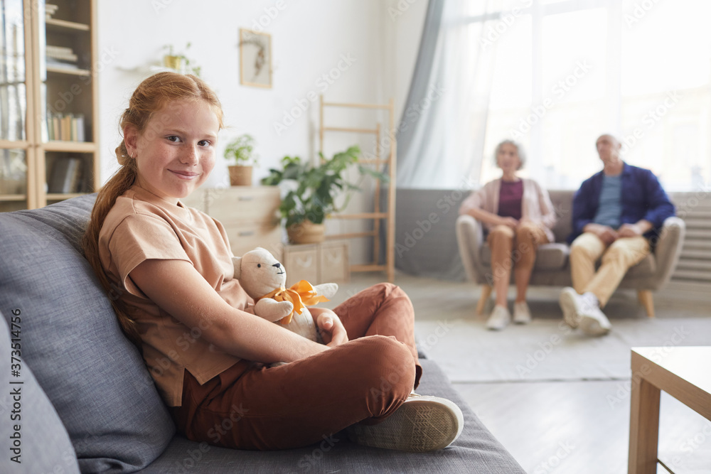 Warm toned portrait of cute red haired girl looking at camera while sitting on couch in cozy living room with grandparents in background, copy space Warm toned portrait of cute red haired girl looking at camera while sitting on couch in cozy living room with grandparents in background, copy space