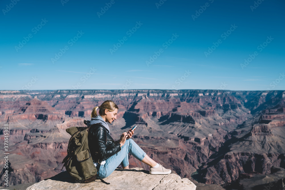 Fototapeta premium Smiling caucasian female travel blogger with backpack using connection on mobile phone for blogging during wanderlust tour in mountains,millennial woman explorer satisfied with connection on cellular