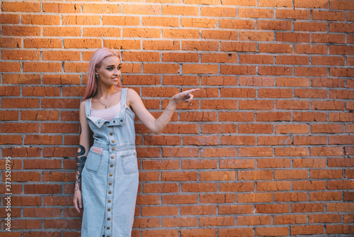 Trendy adolescent teenager dressed in casual jeans sundress smiling and showing something near brick promotional background, happy Caucasian hipster girl pointing near copy space publicity area