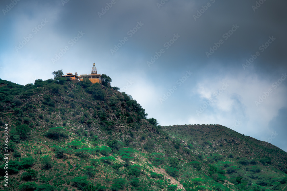 hindu temple on a hill covered in vegetation with the tower and spire clearly visible in the distance with a background of dark monsoon clouds shot in Rajasthan India