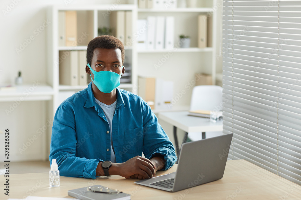 Portrait of young African-American man wearing mask and looking at camera while working at desk in post pandemic office, copy space Portrait of young African-American man wearing mask and looking at camera while working at desk in post pandemic office, copy space