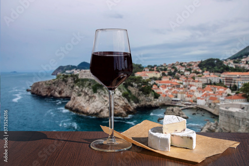 Fototapeta Naklejka Na Ścianę i Meble -  Glass of wine with brie cheese against view of blue sea and rocky coast background. Dubrovnik old town surrounded by old walls. View from above of red rooftops, roofs and fortress.
