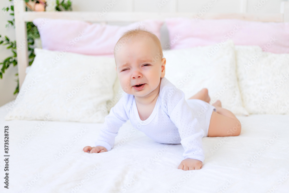 sad baby girl 6 months old in a white bodysuit lying on her stomach on a white bed at home sad baby girl 6 months old in a white bodysuit lying on her stomach on a white bed at home