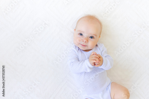 portrait of a small baby girl 6 months old in a white bodysuit lying on her back on a white bed, space for text