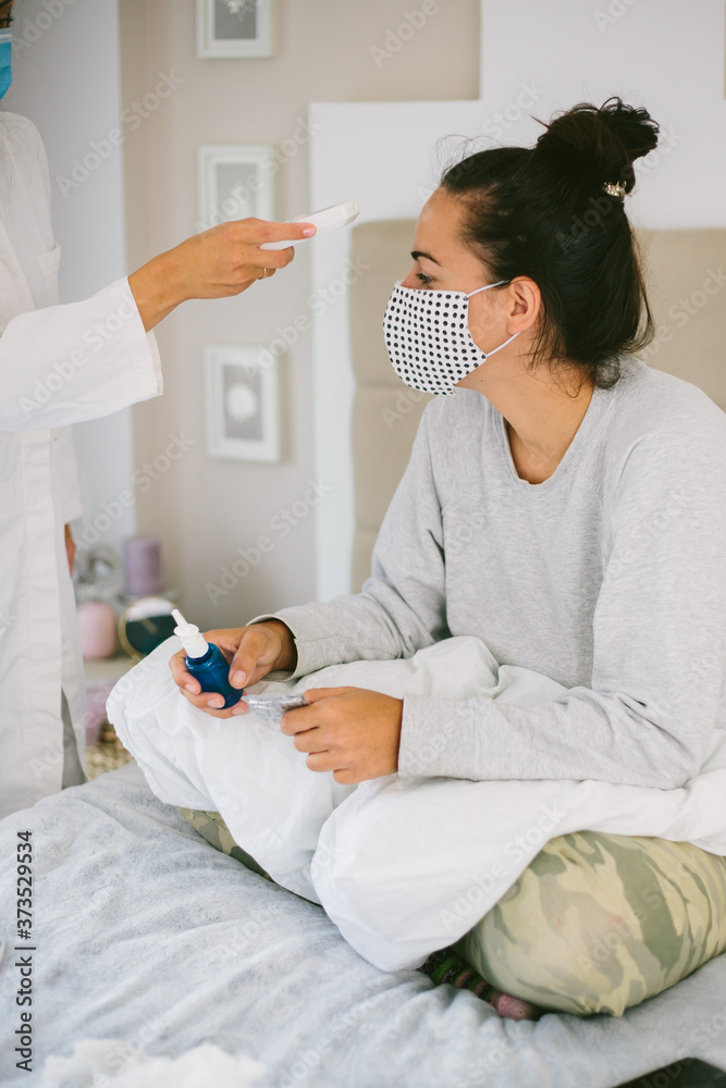Female with protection mask getting her fever checked by a nurse with with mask and a digital thermometer at home.