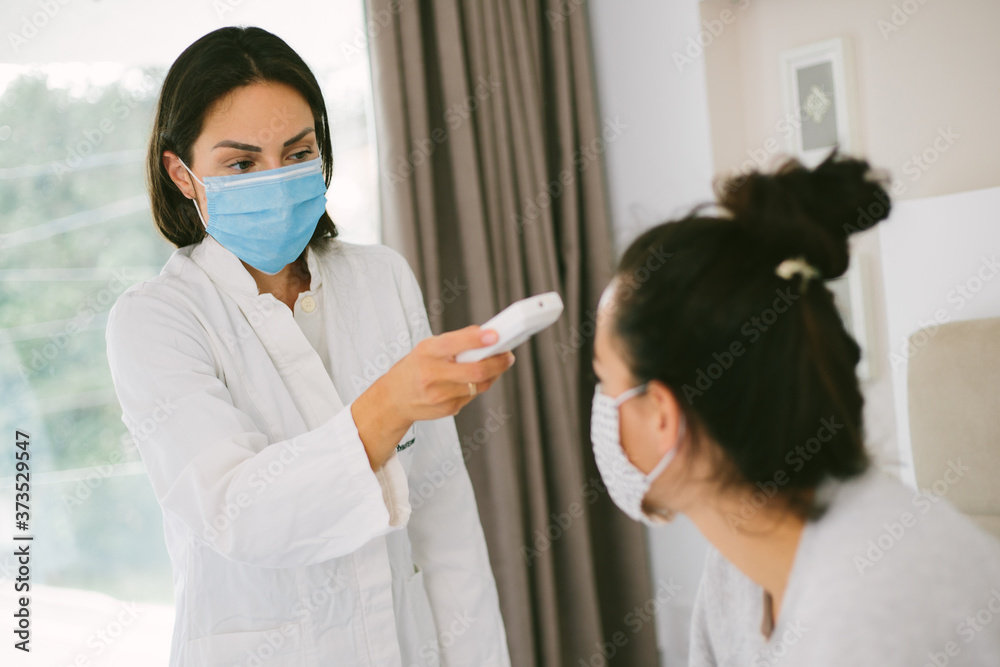 Female with protection mask getting her fever checked by a nurse with with mask and a digital thermometer at home. Female with protection mask getting her fever checked by a nurse with with mask and a digital thermometer at home.