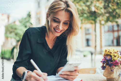 Cheerful female blogger using textbook for planning publication posting sitting in sidewalk and reading notification from social networks, happy hipster girl watching streams video on cellphone