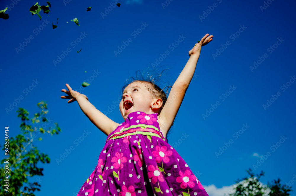 Little girl on the background of beautiful nature. The child threw up flowers against the blue sky. Happy kid portrait photo. Children's smile. The child raised his hands to the sky Little girl on the background of beautiful nature. The child threw up flowers against the blue sky. Happy kid portrait photo. Children's smile. The child raised his hands to the sky