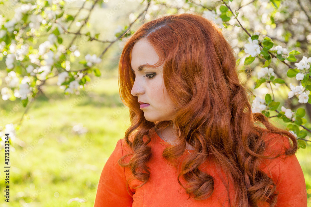 Naklejka premium Portrait of a red-haired girl walking in an apple orchard in an orange dress