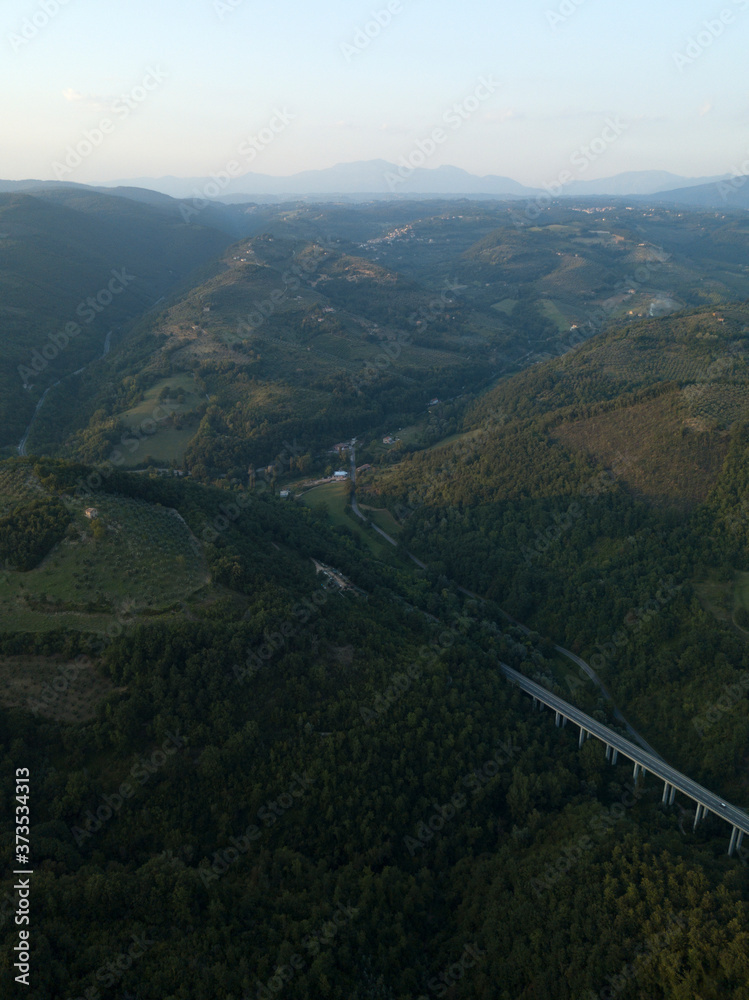 Aerial view of highway road in middle of forest trees on mountains, meadows, italian village Frasso Sabino, Lazio region, Italy, Europe. Green rural landscape countryside. Cars and trucks go on road.