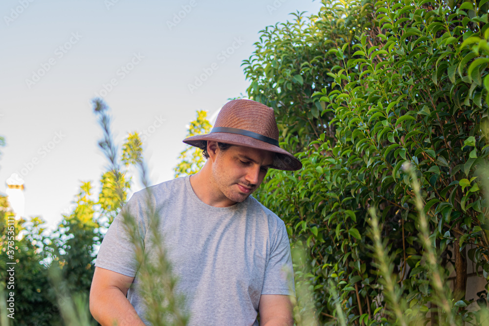Photo of a young and attractive man with a brown hat watering his garden with a watering can during a sunny day. Beautiful flowers, green environment