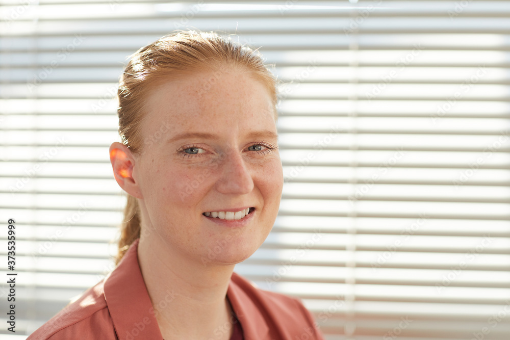 Candid close up portrait of young red haired woman smiling at camera while posing against blinds in sunlit office, copy space Candid close up portrait of young red haired woman smiling at camera while posing against blinds in sunlit office, copy space