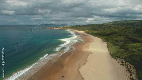 Northern Ireland aerial view: White beach gulf water crashing. People on shore walk and playing with dogs. Ocean and meadows in horizon. Picturesque beauty
