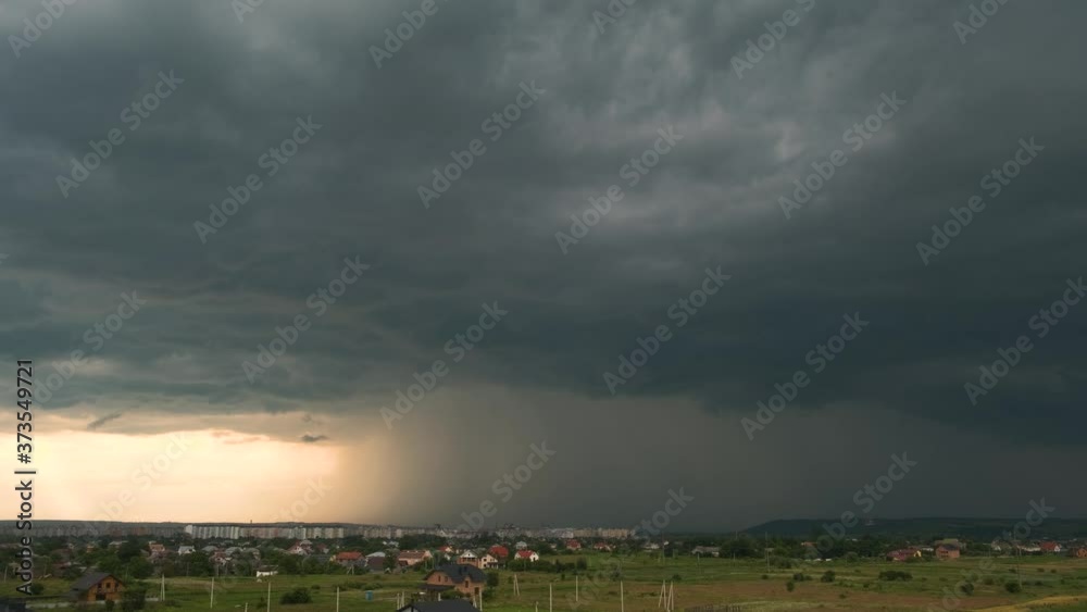 Moody landscape with dark stormy clouds with falling heavy downpour shower rain over distant town buildings in summer.