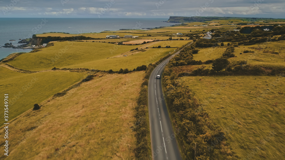 Aerial autumn road: car travel view. Beautiful Northern Ireland countryside nature scene with roadway against Atlantic cliffy shoreline in yellow fall tones