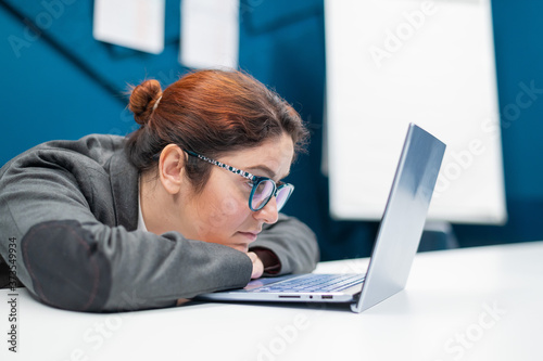 Tired business woman laid her head down on desk and looks at the laptop monitor. Female employee is tired at work in the office.