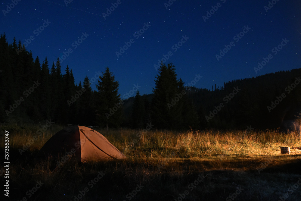 Night photo of a tent near the fire in a camping place. The tent is in a meadow surrounded by coniferous forests. Stars and constellations can be seen on the dark sky. Carpathia, Romania.