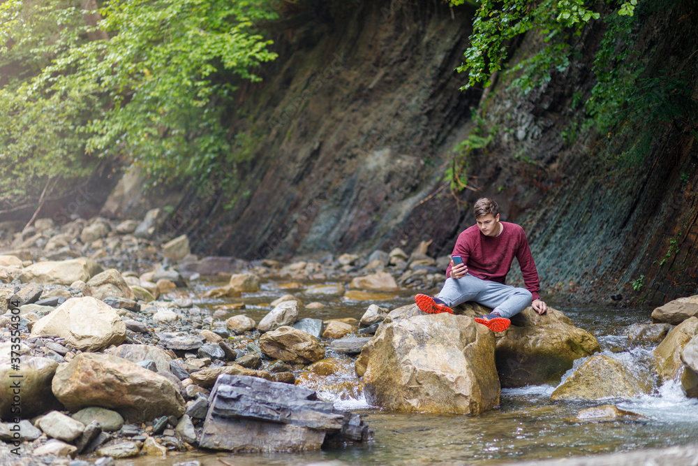 A man looks at the phone while sitting on a stone on a mountain river. A man looks at the phone while sitting on a stone on a mountain river.