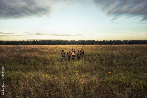 Fototapeta Group of hunters going through rural field towards forest against sunrise sky an
