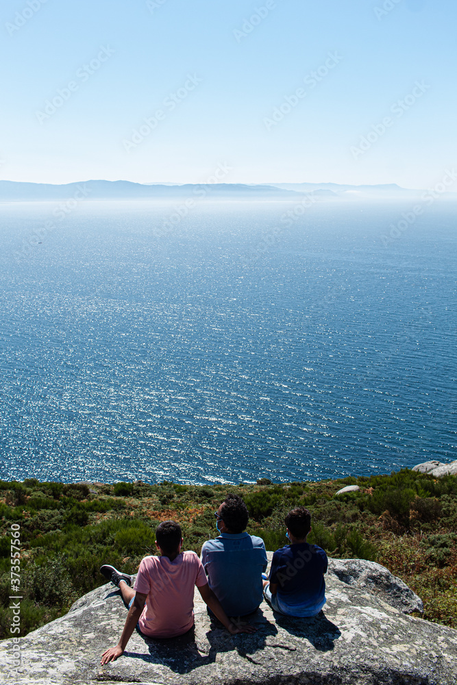 A father with his sons enjoying the breeze and witnessing the Atlantic Ocean while sitting on top of a rock in a viewpoint in the north of Spain