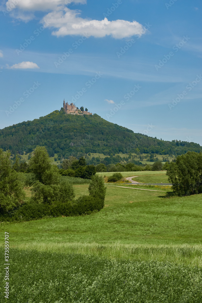 Popular Hohen Zollern castle on a big Hill, green meadow are in the foreground, cyan sky with white Clouds. Germany, Zimmern.