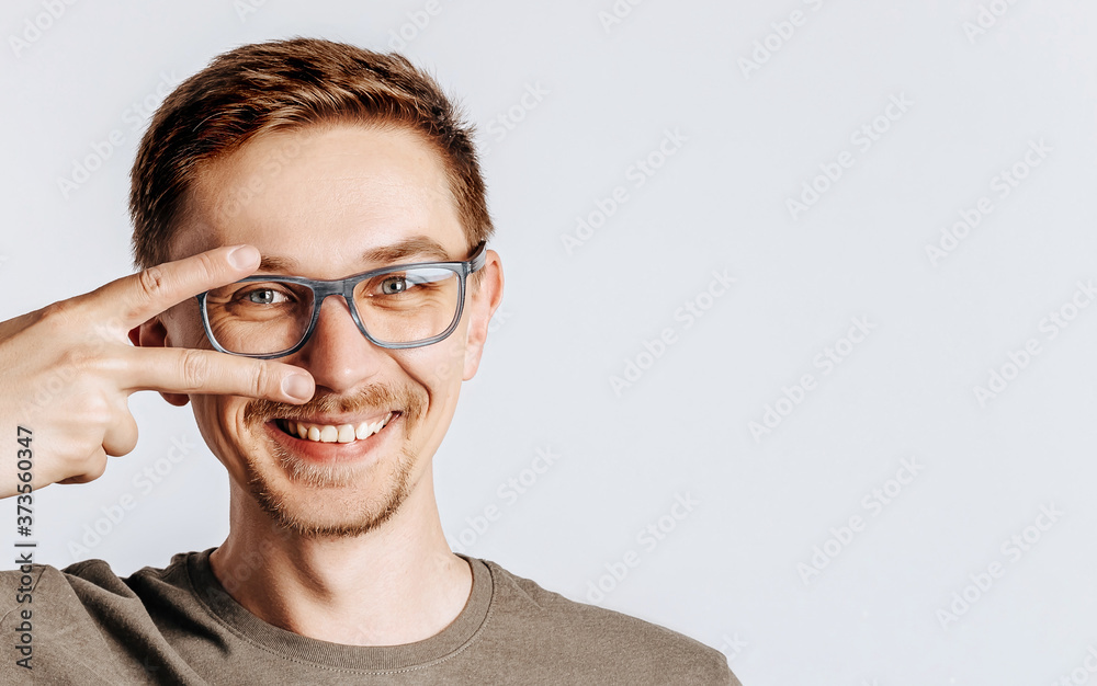 Young handsome man with glasses doing peace symbol with fingers over face, smiling cheerful showing victory. Handsome guy smiles over isolated gray background. Place for advertising.