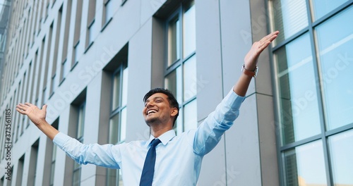 Portrait shot of Hindu young handsome cheerful satisfied man in tie rising hands up to sky and smiling outdoor at business center building. Businessman thankful for success. Successful and rich.