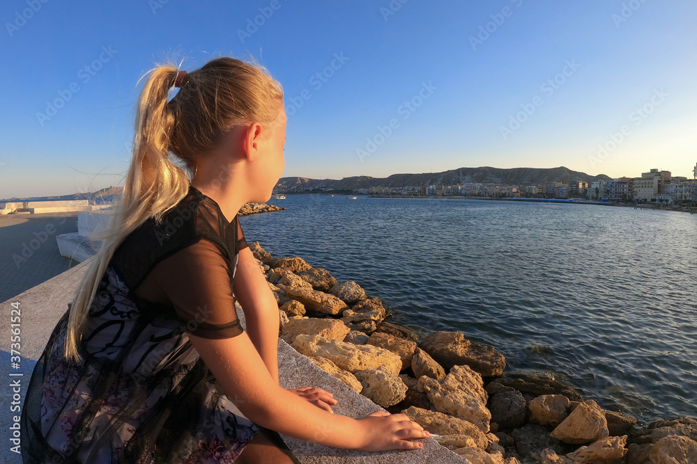 A little blonde girl sits on a sea pier and looks into the distance, a beautiful schoolgirl sits in profile on a white pier and looks at the city beach